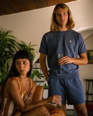 A seated woman in a sleeveless top and a standing man, wearing the matching blue Marseille French Terry Pullover - Moontide by Dandy Del Mar, hold champagne flutes in a room adorned with plants and art.