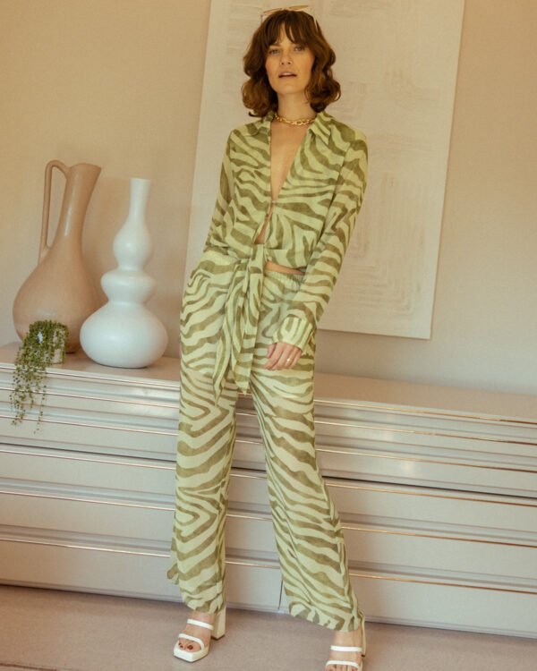 A person stands in a room wearing a vibrant green zebra-patterned outfit paired with white platform sandals, featuring the tie front design of The Cayman Top - Arbequina by Dandy Del Mar, in front of a dresser adorned with decorative vases.