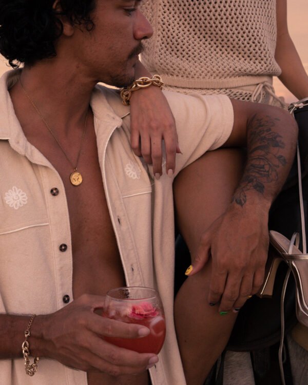 A person with a flower-garnished drink sits near another sporting Dandy Del Mar's stylish The Gaucho Shirt in Alabaster, unveiling a tattooed arm and vibrant, multicolored nails.