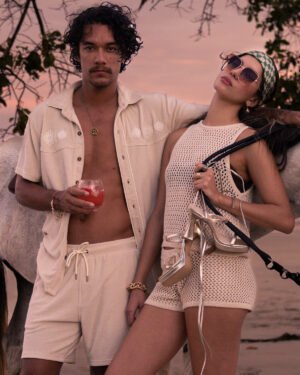 As the sun sets, an Equatorial Cowboy and his companion stand with a white horse. He enjoys a drink, while she, in sunglasses and headscarf, holds her heels. They both wear Dandy Del Mar's The Gaucho Shirt in Alabaster, epitomizing leisurely elegance.