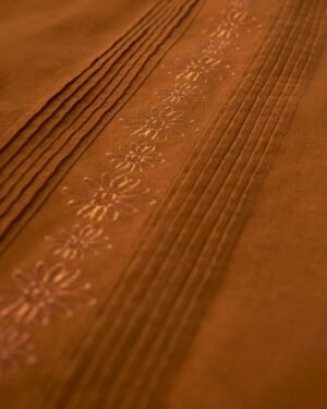 Close-up of an orange-brown Brisa Linen Shirt - Sedona by Dandy Del Mar, featuring gardenia floral embroidery and several parallel pleats, accented by coconut shell buttons.