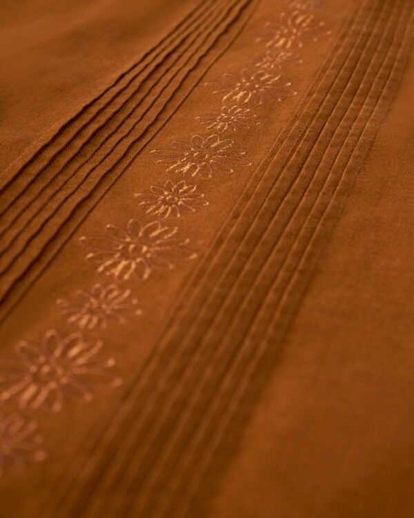 Close-up of an orange-brown Brisa Linen Shirt - Sedona by Dandy Del Mar, featuring gardenia floral embroidery and several parallel pleats, accented by coconut shell buttons.