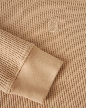 Close-up of a beige, ribbed-texture fabric with a cuffed sleeve made from 100% cotton. An embroidered logo is visible on the main fabric of Dandy Del Mar's Cannes Long Sleeve Shirt in Truffle.