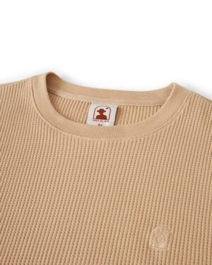 Close-up of The Cannes Long Sleeve Shirt in Truffle by Dandy Del Mar, a beige waffle-knit shirt made from 100% cotton, featuring a small logo emblem on the bottom right and a neck tag reading 