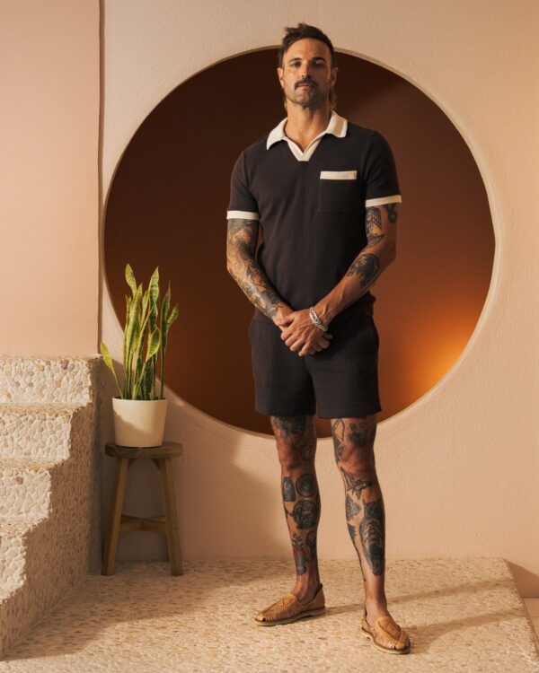 A man with arm and leg tattoos stands in a room, wearing a dark short-sleeved Cannes Waffle Knit Shirt - Carajillo from Dandy Del Mar, paired with shorts. He is posed by a round wall feature and a plant on a stool.