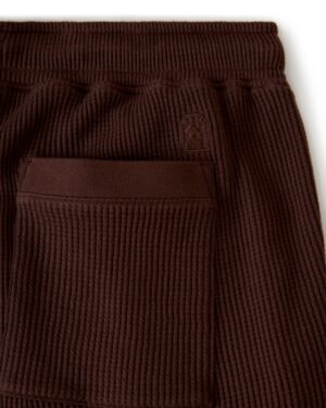 Close-up of dark brown The Cannes Waffle Knit Shorts by Dandy Del Mar, showcasing their ribbed fabric, elastic waistband, and a small embroidered logo above a back pocket.