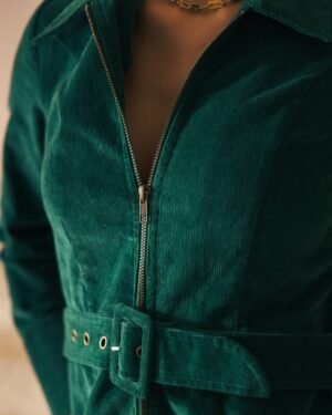 A close-up of a person wearing The Kitts Corduroy Dress - Albero by Dandy Del Mar, showcasing its green corduroy fabric, zipper, and slim-fit waist belt adorned with grommets.