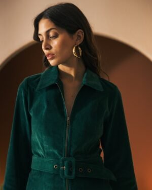 A woman with long hair, adorned with earrings, is looking down while wearing The Kitts Corduroy Dress - Albero by Dandy Del Mar. The slim-fit green zip-up dress features a belted waist and she stands against an arch backdrop.