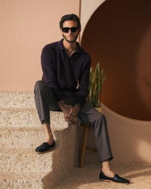 A man in sunglasses and The Sebastian Long Sleeve Polo by Dandy Del Mar sits on the stairs with a potted plant nearby, all against a light pink and brown circular wall backdrop.