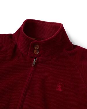 A close-up of the sangria red Italian corduroy Corsica Corduroy Jacket by Dandy Del Mar, featuring a high collar, zipper, and two brown buttons. The jacket showcases an embroidered logo on the left side.