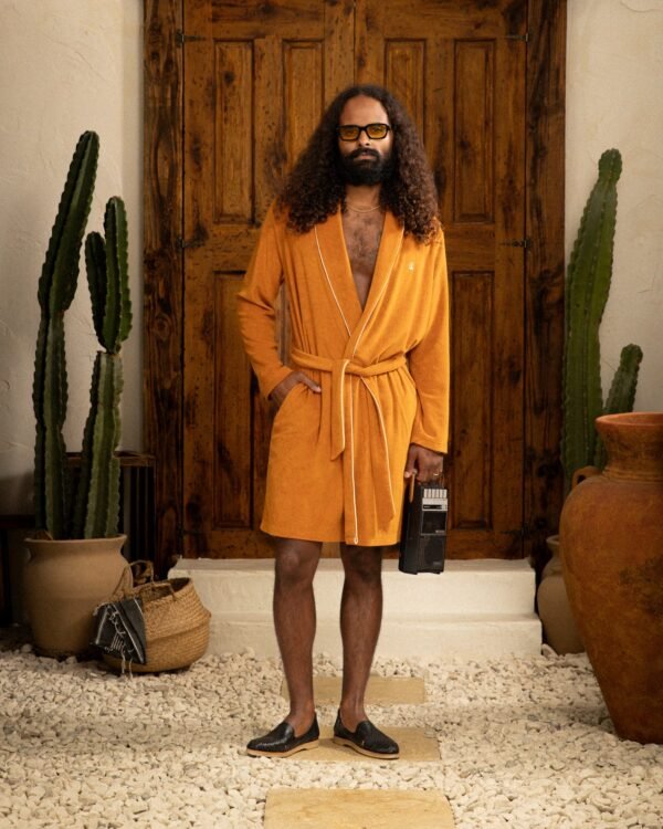 A person with long hair and glasses, wearing the above-the-knee "The Tropez Robe - Burnt Sienna" by Dandy Del Mar, stands in black shoes holding a boombox. The stone path is lined with cacti, and a wooden door serves as their backdrop.