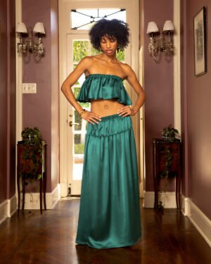 A person is wearing a teal, off-the-shoulder, ruffled top paired with The Monaco Maxi Skirt in Albero by Dandy Del Mar, standing in a hallway with purple walls and wooden flooring.