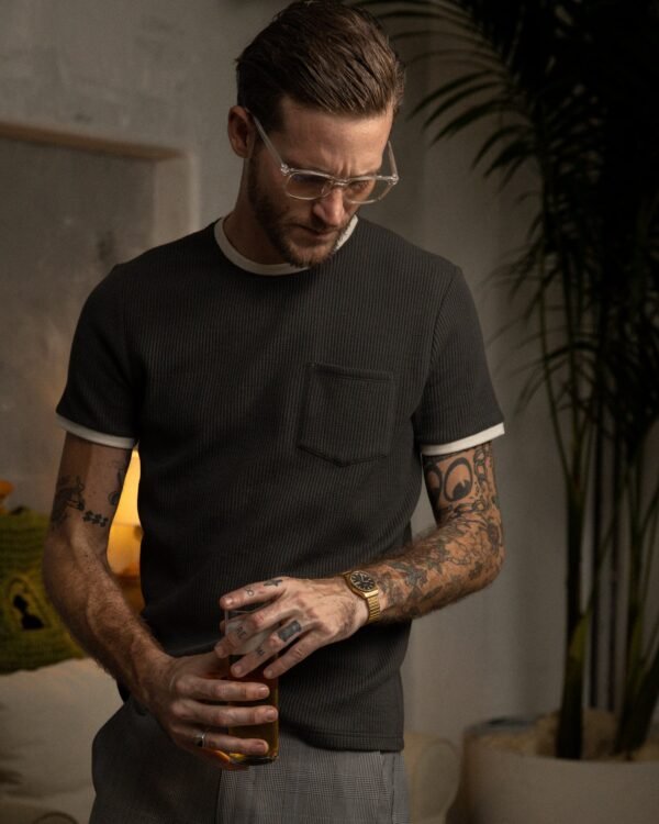 A tattooed man in glasses and a dark, breathable "The Cannes Waffle Knit Tee - Albatross" by Dandy Del Mar holds a drink in a dimly lit room with plants and a couch behind him.