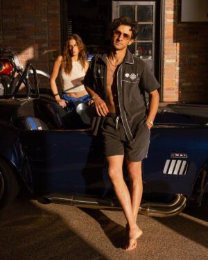 A man in the Onyx Gaucho Shirt by Dandy Del Mar leans against a dark sports car near a garage, projecting an equatorial cowboy vibe. Nearby, a woman in a beige terry cloth top stands, with part of a motorcycle visible in the background.