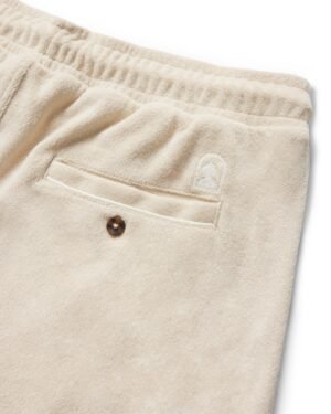 Close-up of the terrycloth shorts in beige from Dandy Del Mar, known as The Gaucho Shorts - Alabaster. They feature an elastic waistband, buttoned back pocket, and a logo above it. Ideal for capturing the Equatorial Cowboy vibe with a modern twist.